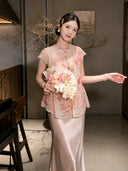 Front view of a pink sheer embroidered vest with a ruffled hem, styled with a matching satin skirt and holding a floral fan.
