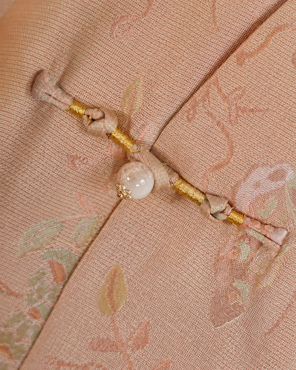 Close-up of a handmade frog closure with gold thread and a pearl-style accent on blush pink silk