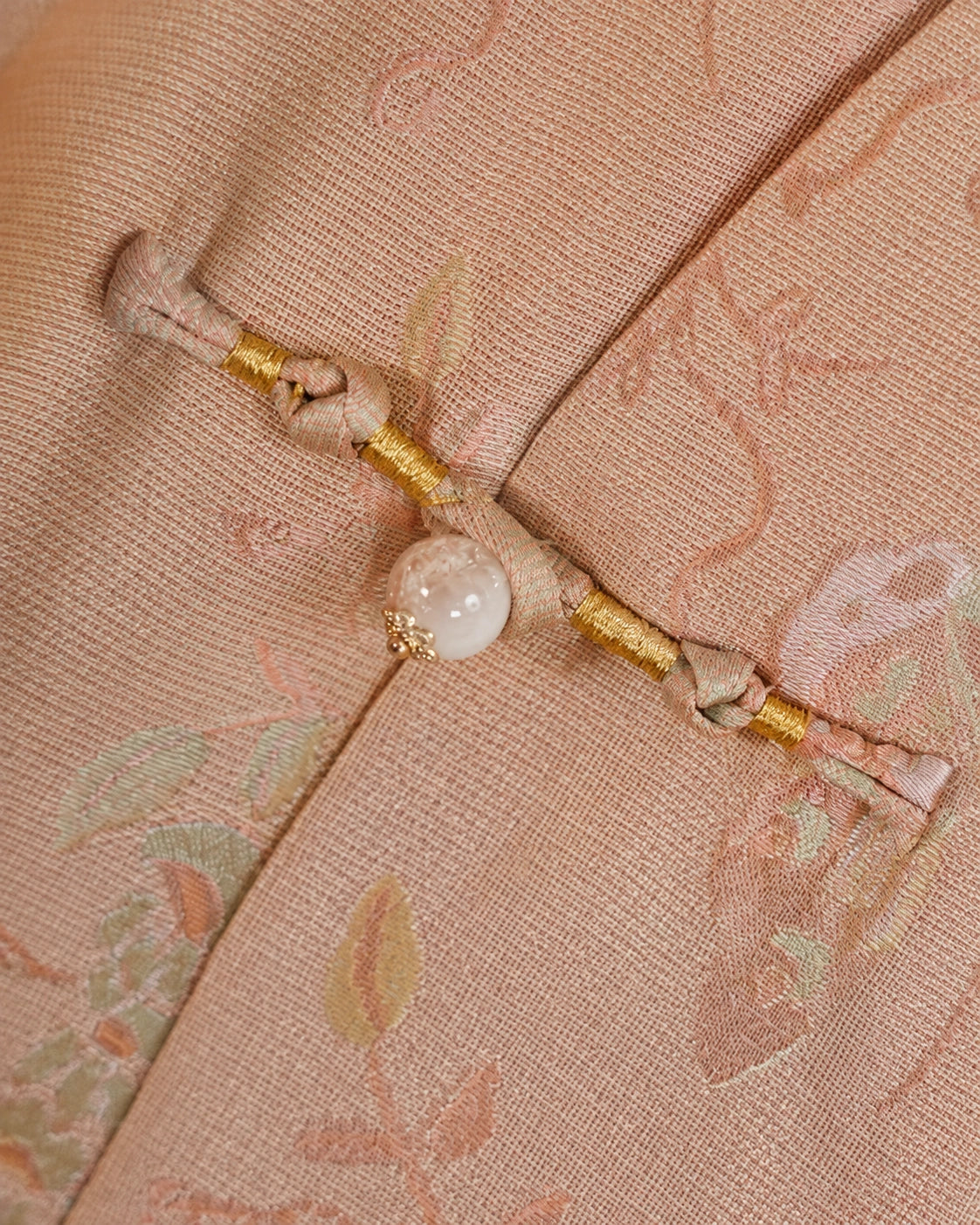 Close-up of a handmade frog closure with gold thread and a pearl-style accent on blush pink silk