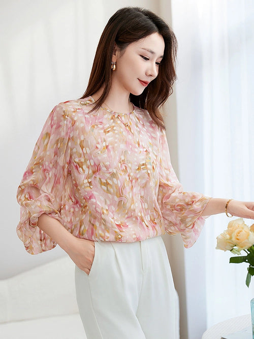 Model wearing a pink abstract printed silk blouse with lantern sleeves and a round neckline, paired with white trousers, showcasing the relaxed and flowy fit.