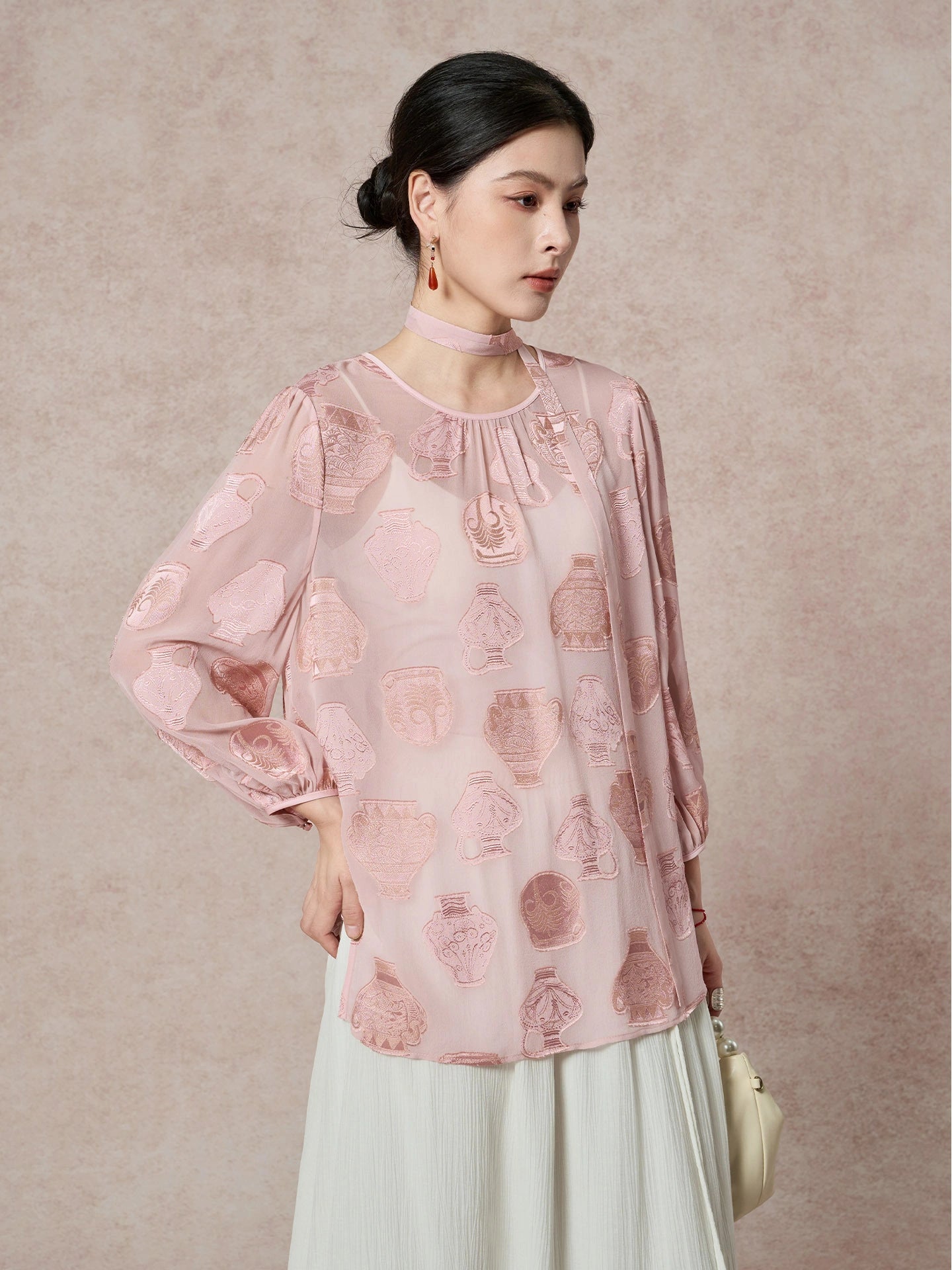 Model wearing a pink sheer silk blouse with an ancient pottery cutwork pattern and a matching necktie, paired with a cream skirt.