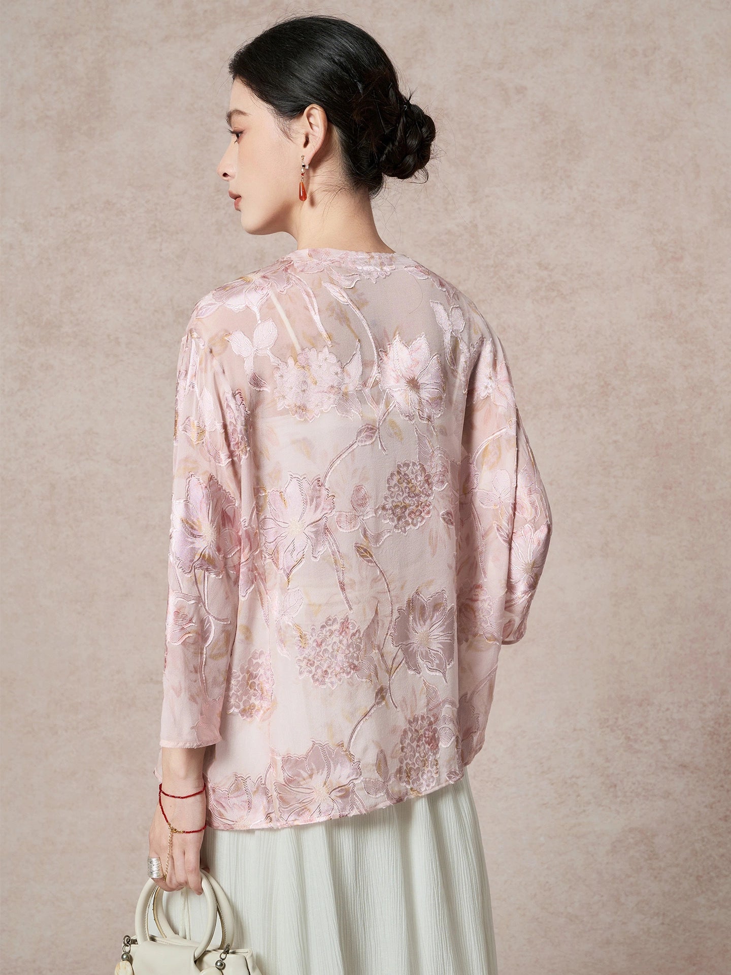 Back view of a sheer pink 100% silk blouse featuring delicate cutwork floral embroidery and a round neckline, paired with a pleated skirt.