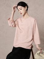 Model seated in a pink 93.8% mulberry silk blouse with a round neck, diagonal placket, and intricate jacquard patterns, styled with a black skirt.