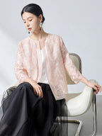Model wearing a pink sheer silk organza embroidered cardigan over a white camisole, sitting on a chair, showcasing the lightweight and elegant layering piece.