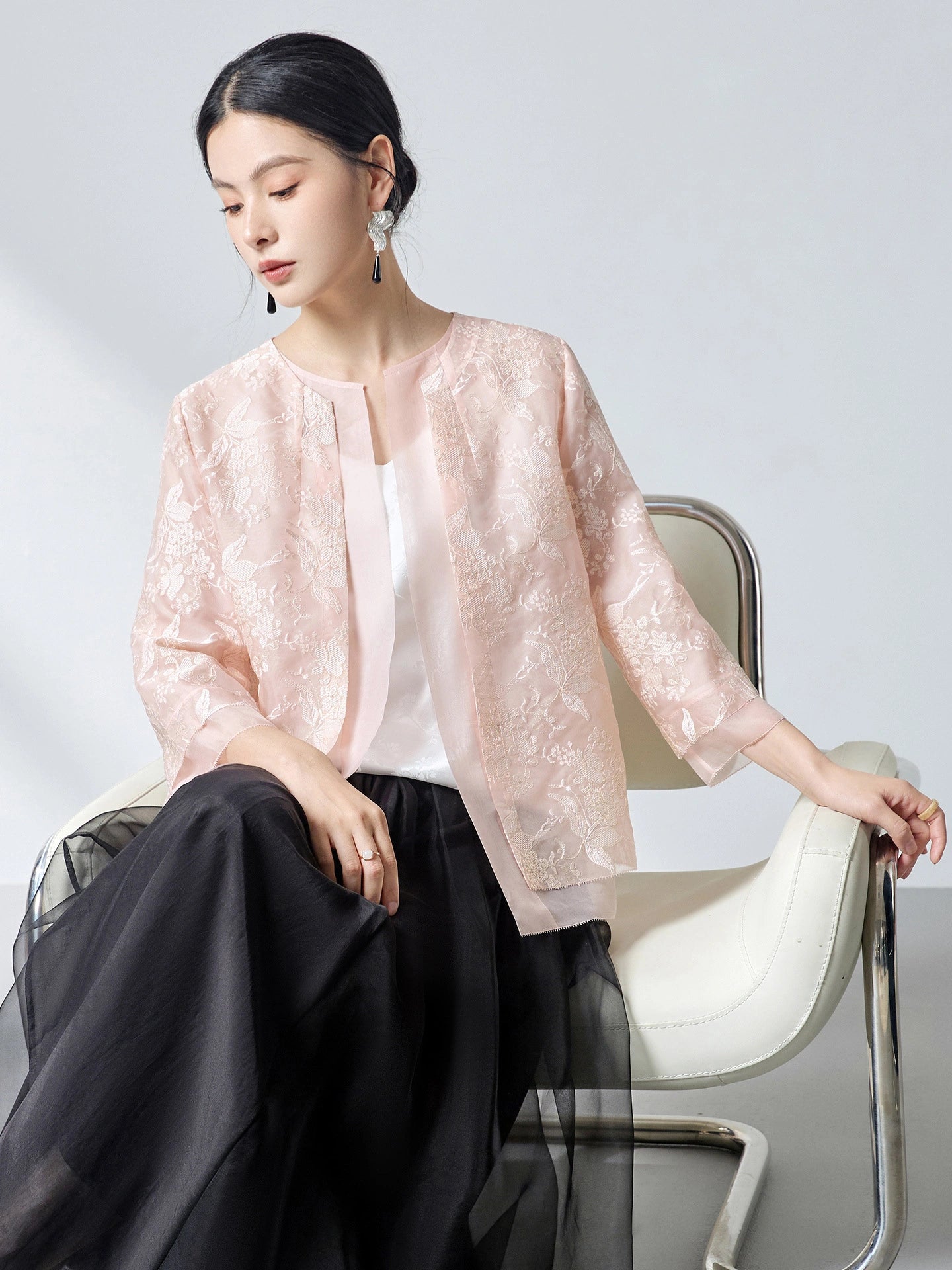Model wearing a pink sheer silk organza embroidered cardigan over a white camisole, sitting on a chair, showcasing the lightweight and elegant layering piece.