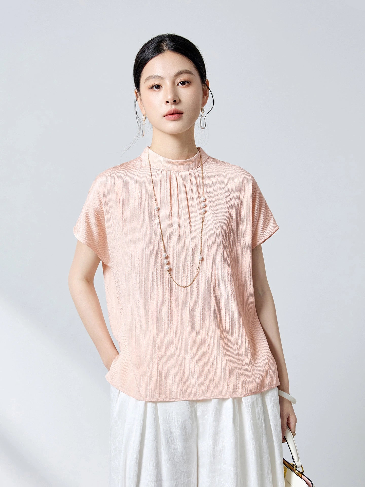 Front view of a pink short-sleeve silk top featuring a stand collar, delicate front pleats, and a relaxed fit, paired with white pleated pants.