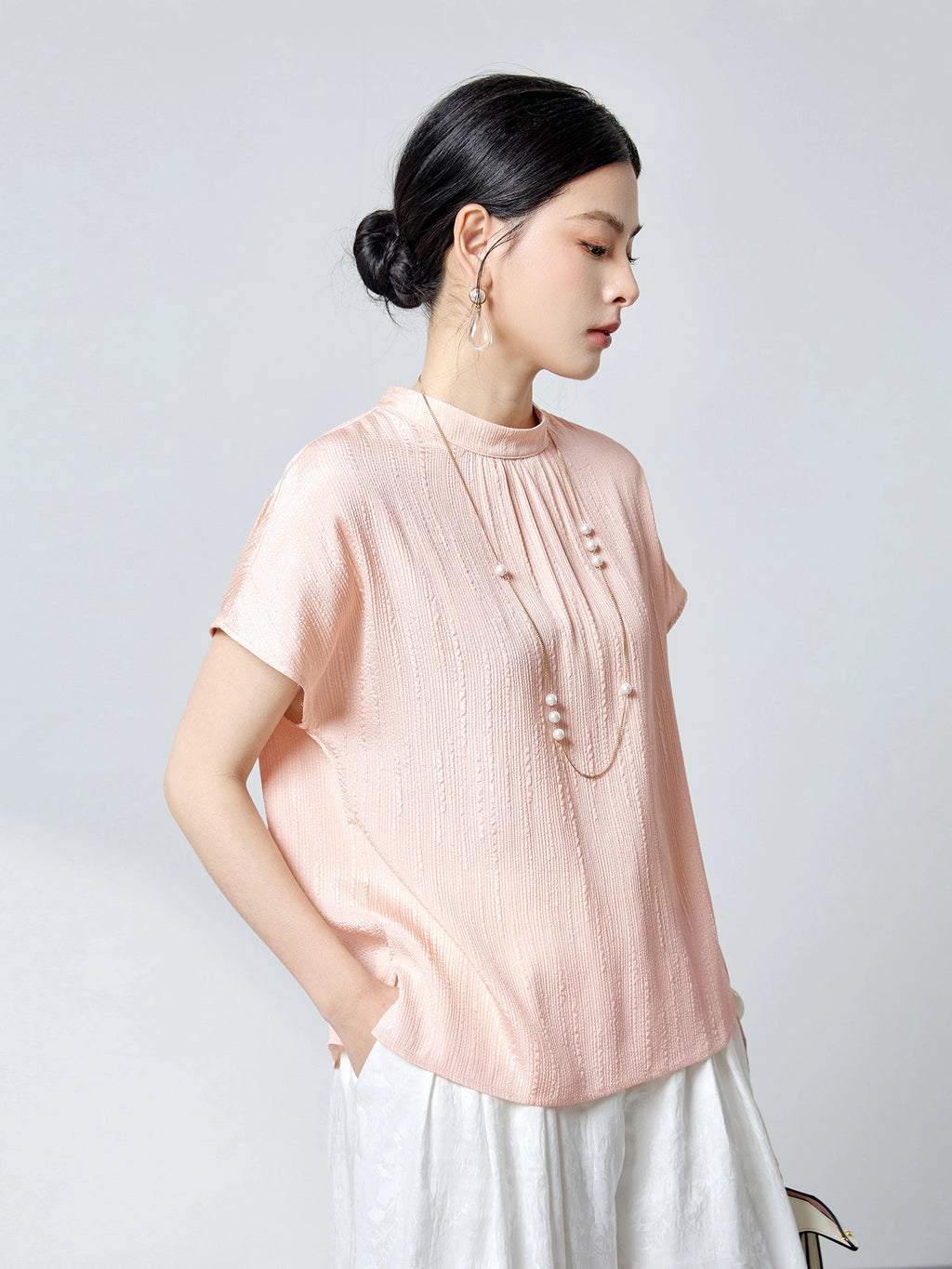 Side view of a pink silk blouse with a stand collar and subtle textured pleats, styled with a long pearl necklace, highlighting the elegant drape and silky sheen.