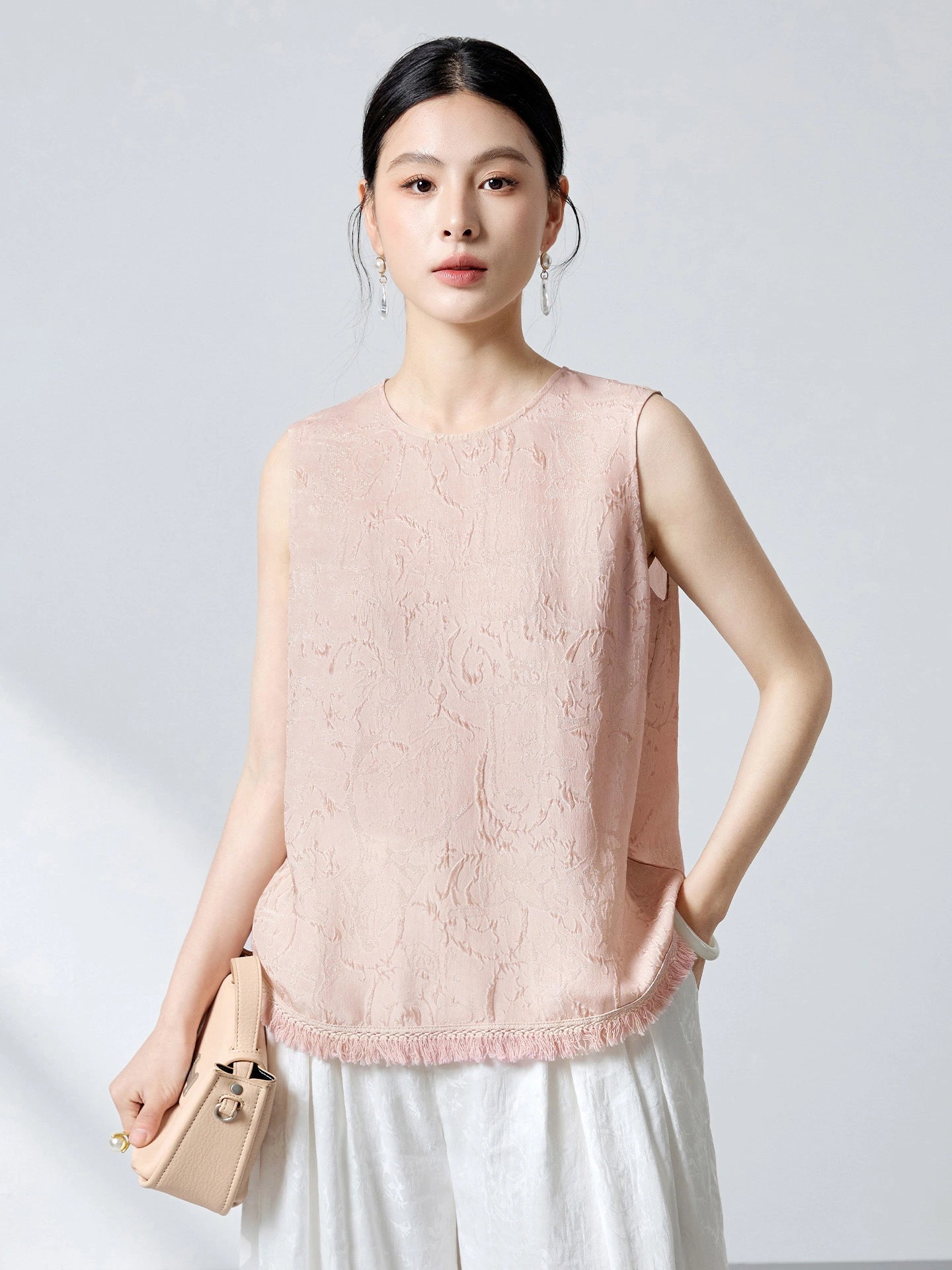 Front view of a soft pink sleeveless silk top with a round neckline, floral jacquard pattern, and tassel hem, paired with white pants.