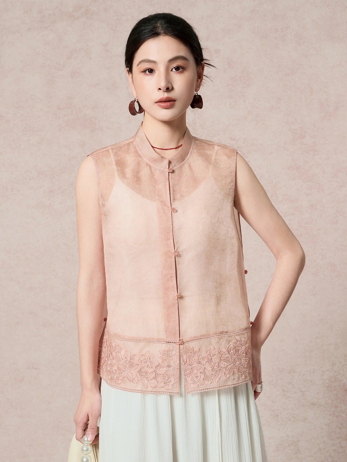 Model wearing a pink 100% silk organza vest with floral embroidery, holding a bag, paired with a white pleated skirt.