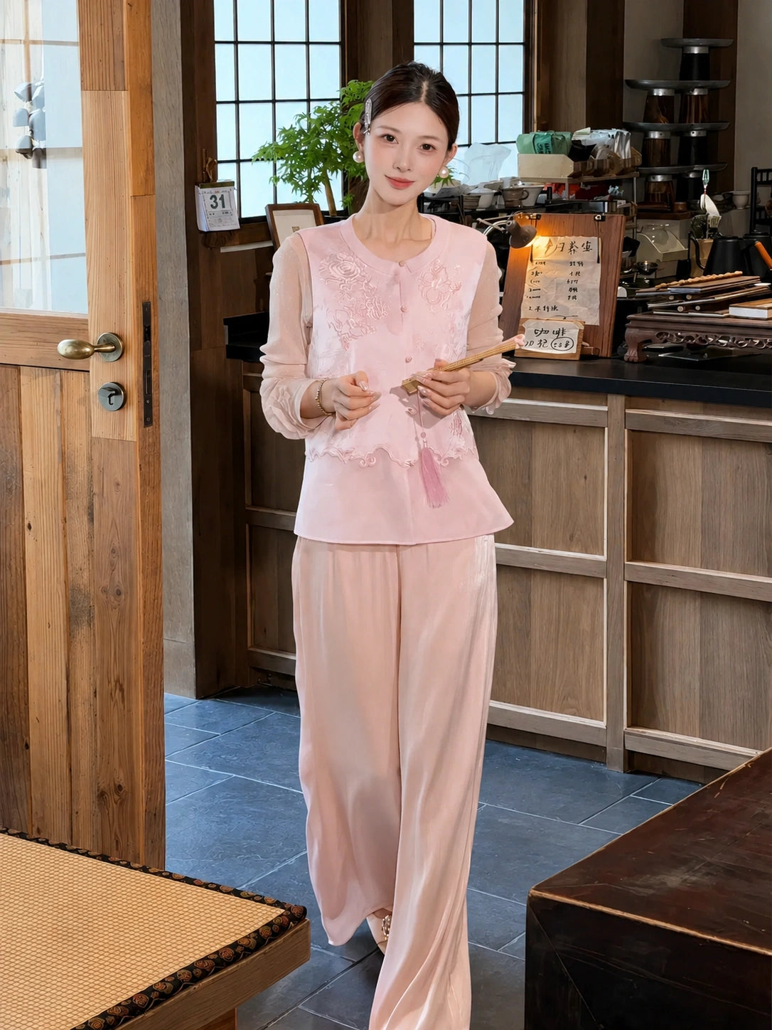 Model in a pink embroidered top and matching pink wide-leg pants, holding a fan in a tea room setting.