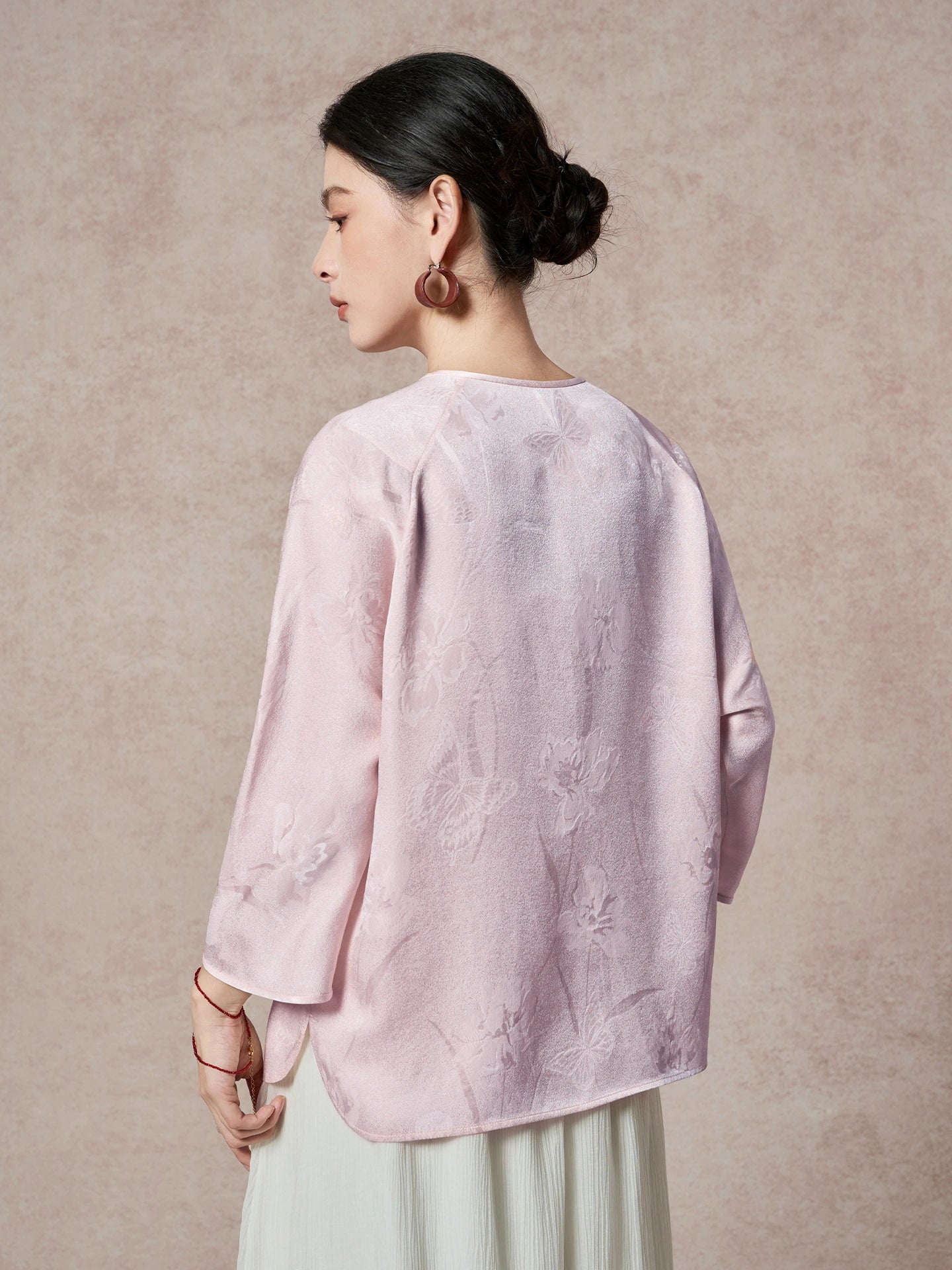 Back view of a purple silk blouse featuring a round neck, raglan sleeves, and a subtle floral jacquard pattern, paired with a white pleated skirt.