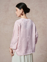 Back view of a purple silk blouse featuring a round neck, raglan sleeves, and a subtle floral jacquard pattern, paired with a white pleated skirt.