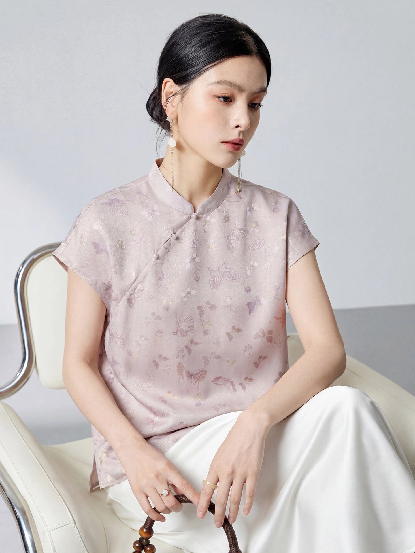 Model seated in a light purple 94% silk top with a stand collar, butterfly jacquard, and pearl buttons, holding a brown handbag, highlighting the elegant drape.