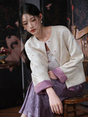 Seated model wearing the ivory side of a reversible floral jacquard jacket with purple turned-back cuffs and a purple skirt.