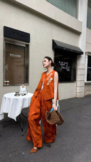 Model standing by stone wall in a rust orange Cheongsam set, looking to the side, with white floral embroidery on the vest.