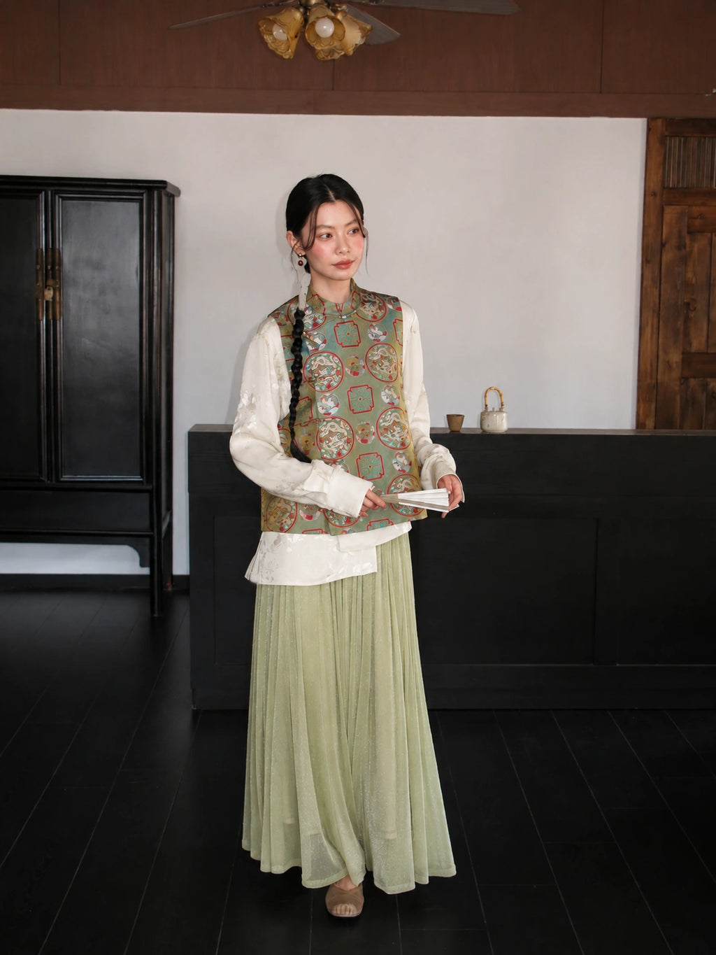 Model wearing a sage green Songjin silk Chinese vest with dragon and phoenix motifs over a white blouse.