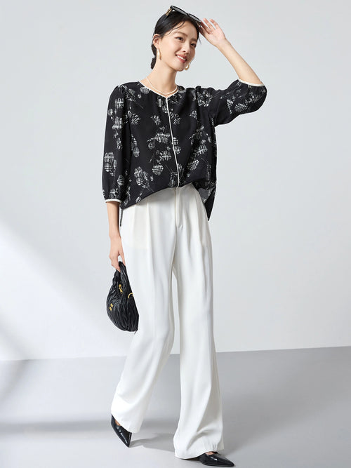 Model wearing a black and white printed 100% silk shirt with puff sleeves and a round neckline, paired with white wide-leg pants, holding a black handbag.