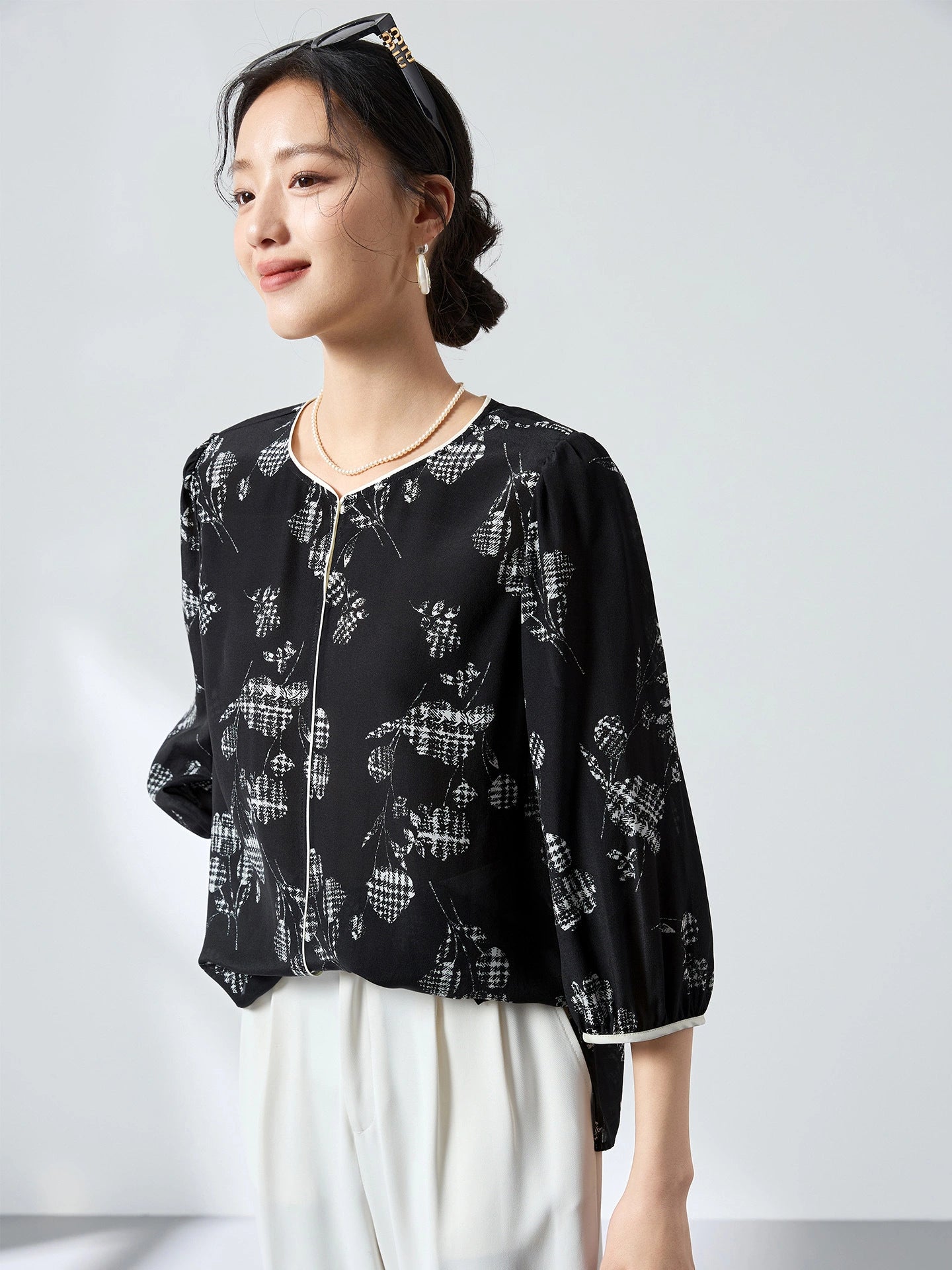Front close-up of a black printed silk shirt featuring a round neckline with cream piping and a relaxed fit, styled with a pearl necklace.