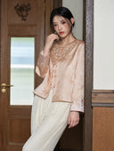 Model leaning by a doorway in the soft peach pink jacket, showing its embroidered neckline and cuffs.