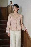Model wearing the soft peach pink embroidered jacket with ivory trousers beside a staircase.