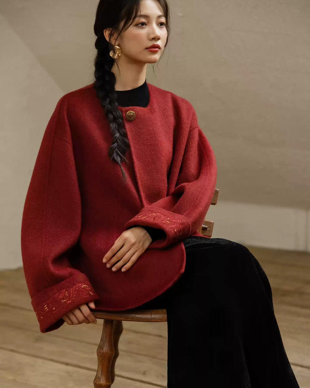 Seated model wearing the red Suian coat, with tonal embroidery and gold-tone accents visible on both cuffs