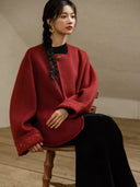 Seated model wearing the red Suian coat, with tonal embroidery and gold-tone accents visible on both cuffs