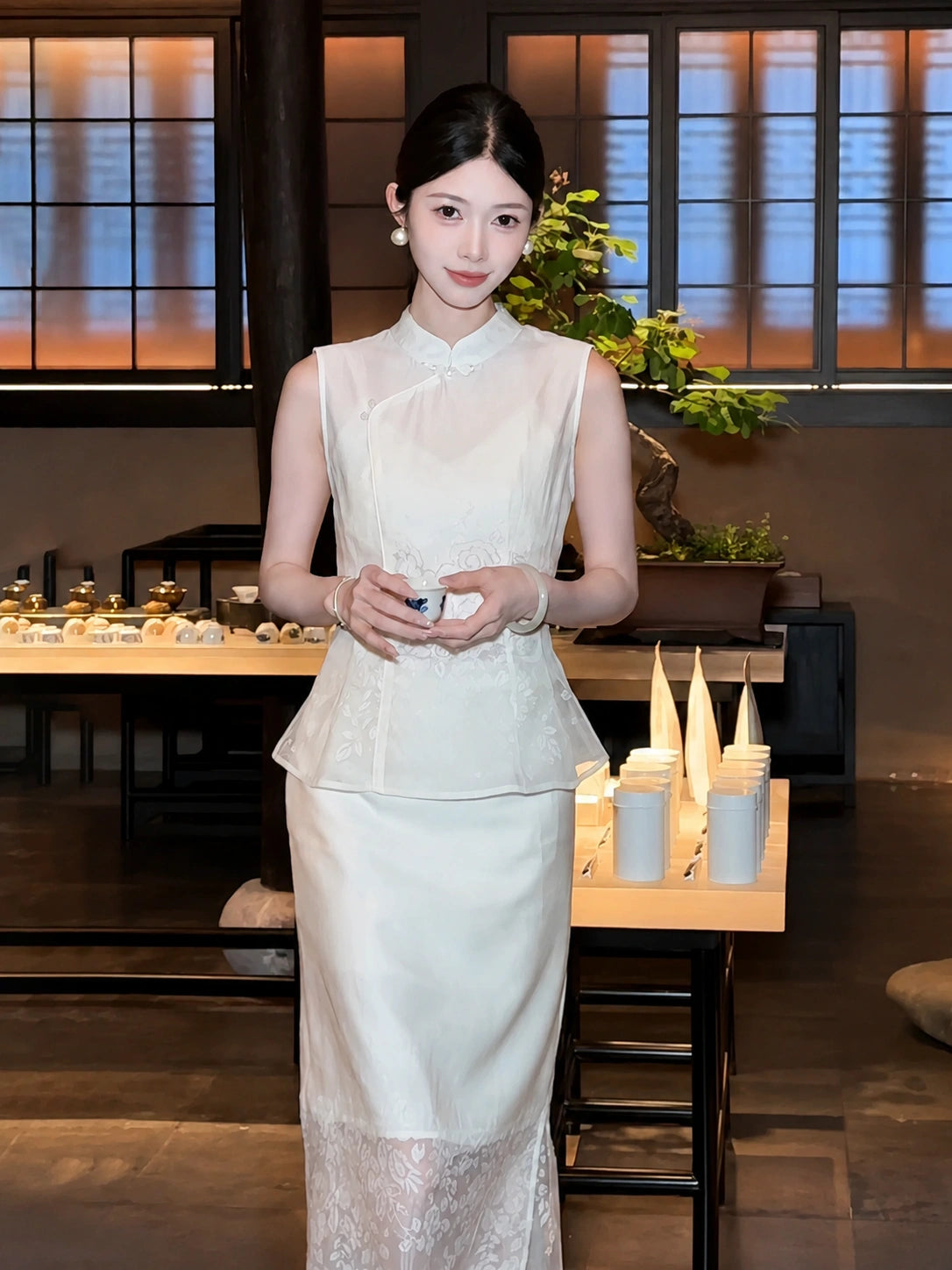 Front view of a white Qipao blouse and A-line skirt set with lace details, worn by a model holding a teacup in a traditional interior.