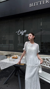 Full body shot of a white Cheongsam blouse and maxi skirt set, featuring a mandarin collar and buttoned front, paired with white heels and a white bag.