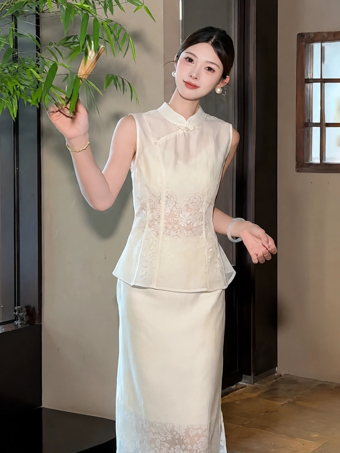 Woman wearing a white sleeveless Cheongsam top and matching skirt with floral embroidery, holding bamboo leaves, featuring a mandarin collar.