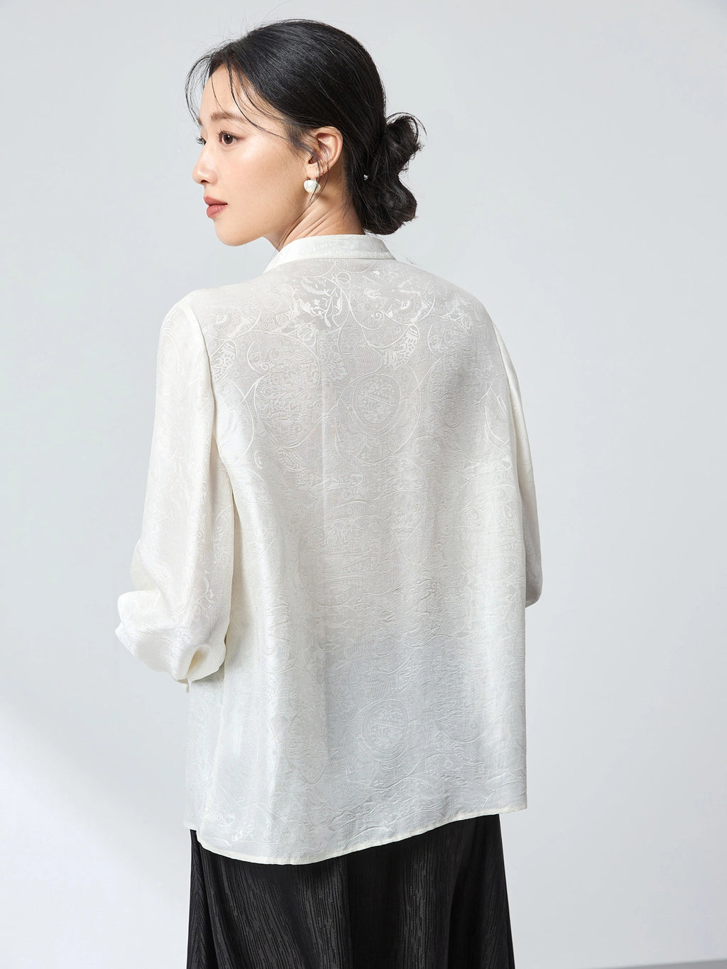 Model wearing a white jacquard blouse with a stand collar, paired with a black pleated skirt, showcasing the flowy silhouette and subtle sheen of the silk-blend fabric.