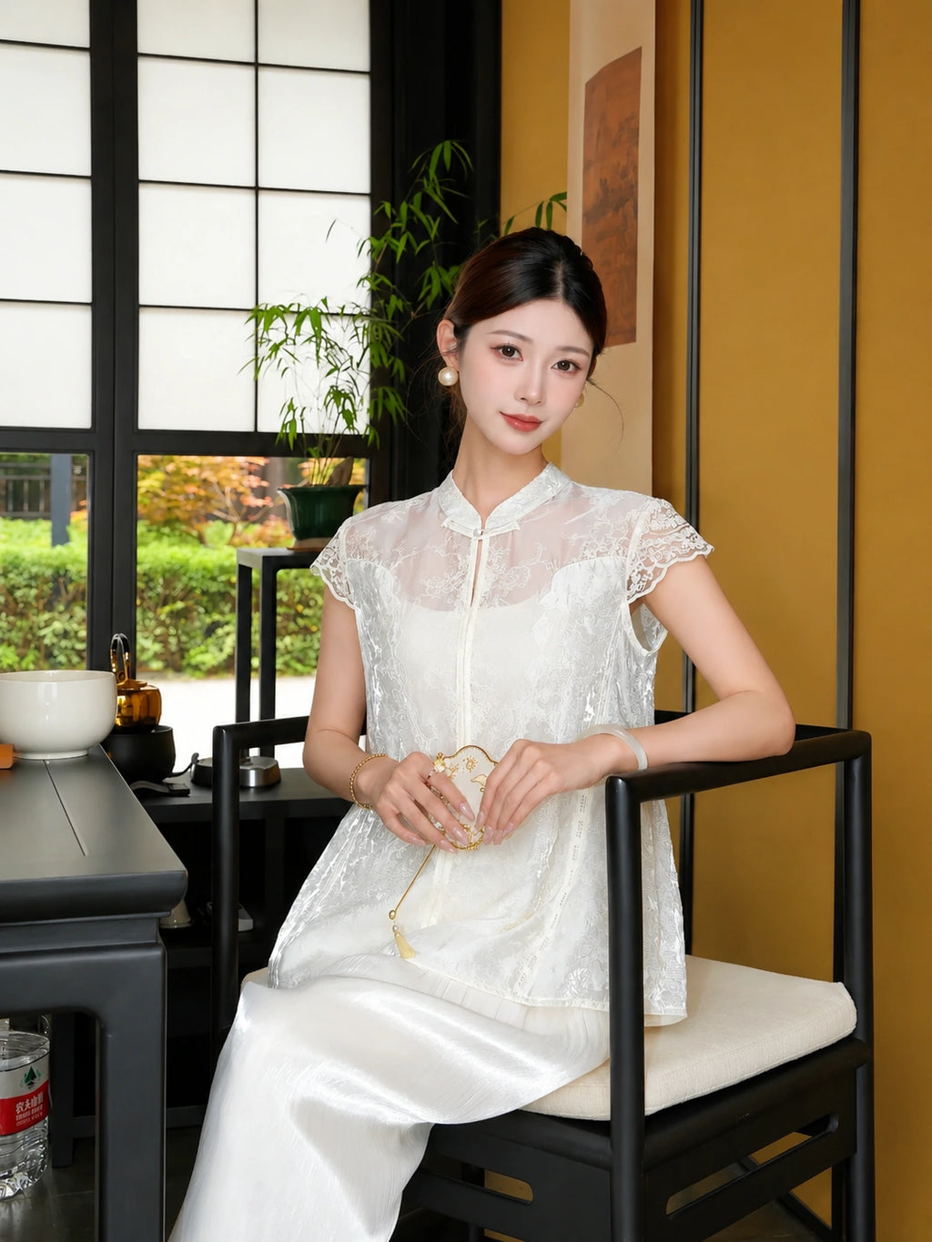 Sitting model in a white lace-trimmed Cheongsam top with a mandarin collar, holding a fan, styled with white pants.