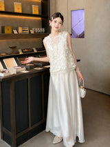 Side view of a white shiny pleated skirt styled with a floral appliqué top and pearl necklace, standing in a boutique setting.