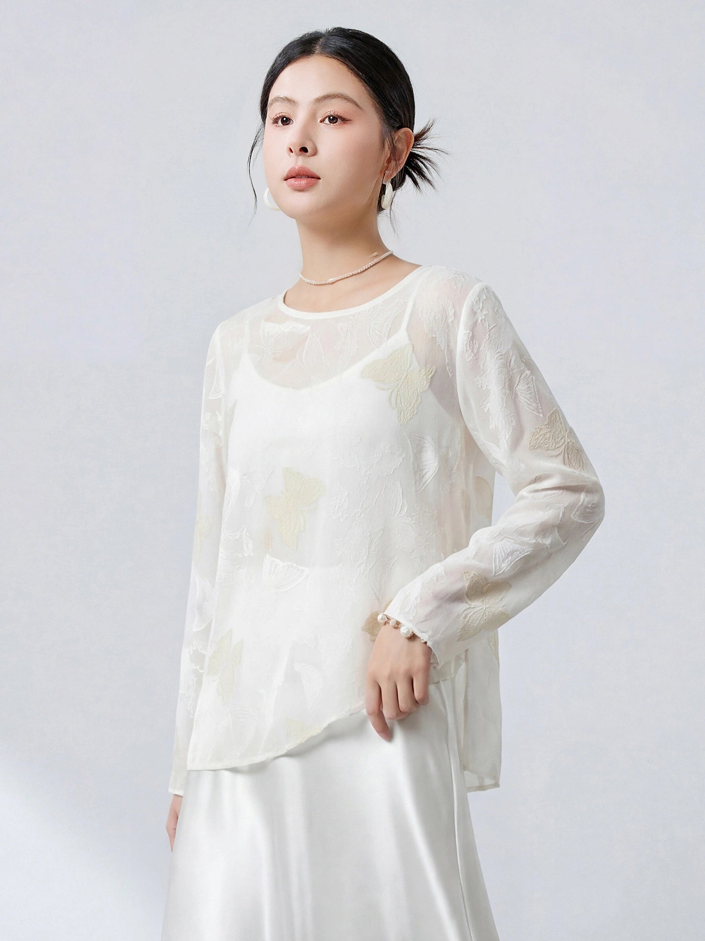 Front view of a white sheer silk top with a relaxed fit, butterfly cutwork embroidery, and a subtle sheen, styled with a white satin skirt.