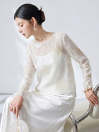 Side view of a model wearing a sheer white 100% silk top with butterfly cutwork embroidery, sitting and holding a round fan, paired with a white satin skirt.