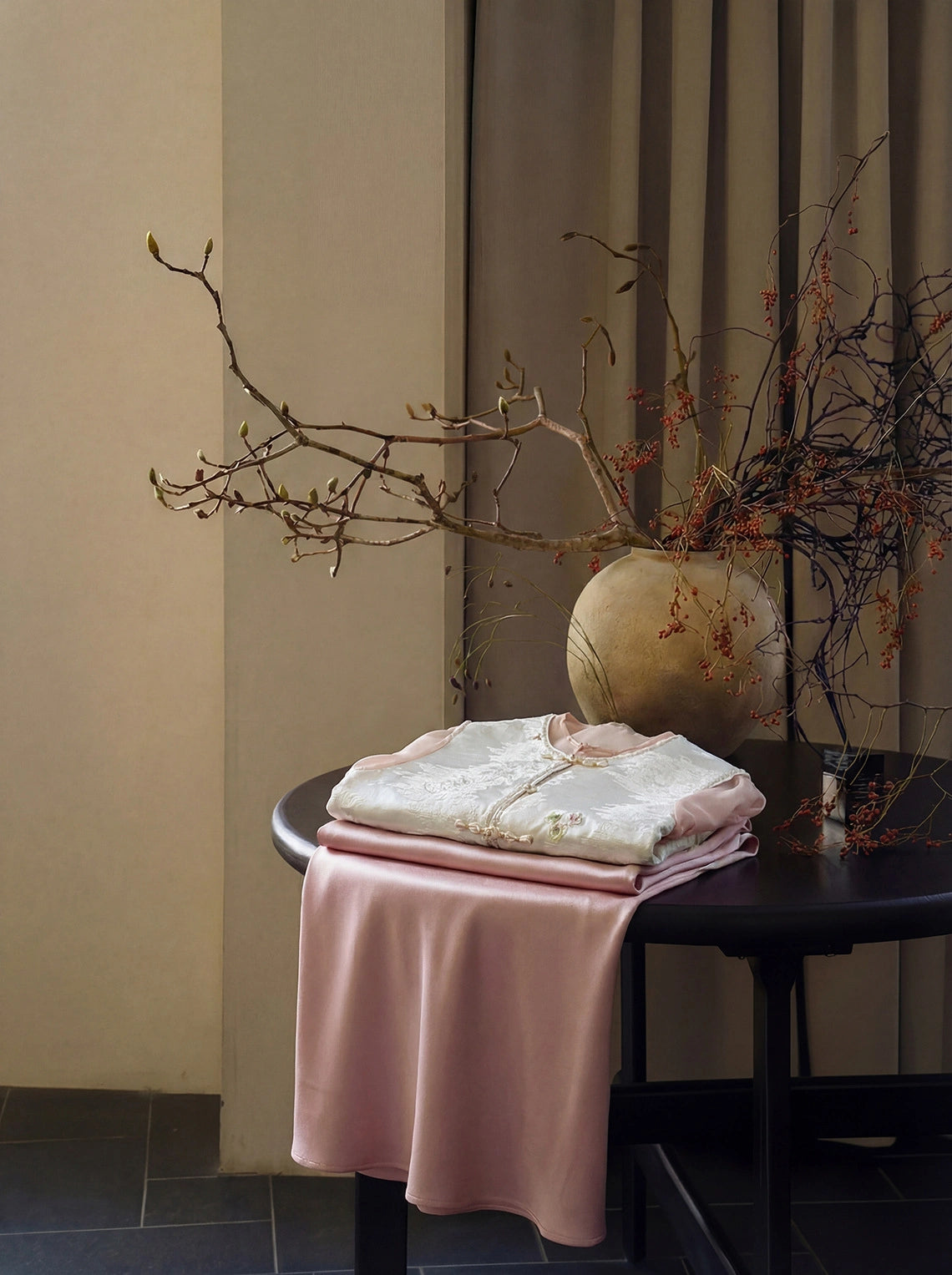 Still life of a white New Chinese style Tencel embroidered vest with traditional frog buttons, styled with a pink satin skirt and dried branches.