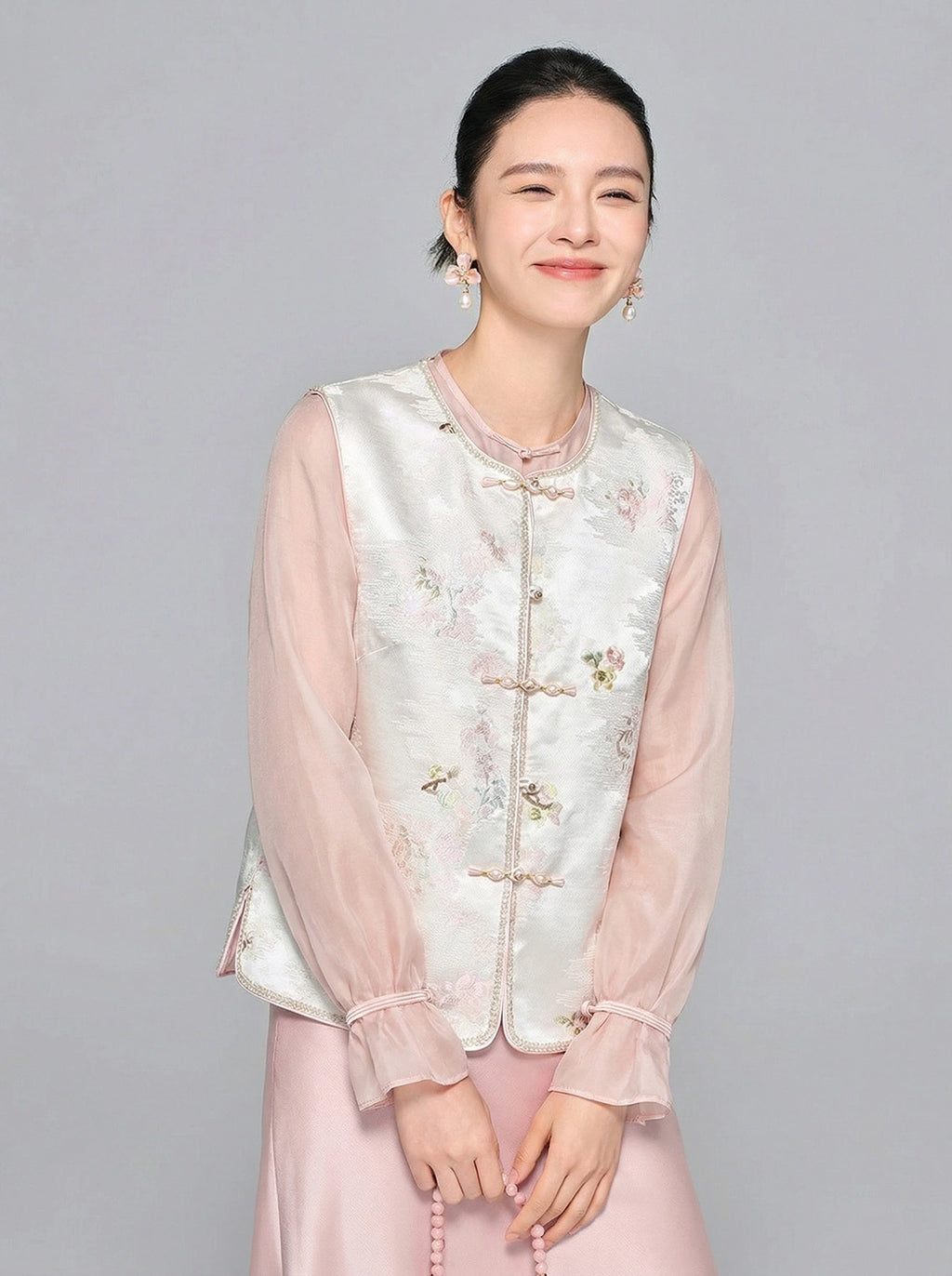 Front close-up of a women's white Tencel vest featuring a round neckline, delicate floral embroidery, and elegant frog buttons, styled with a pink sheer top and satin skirt.