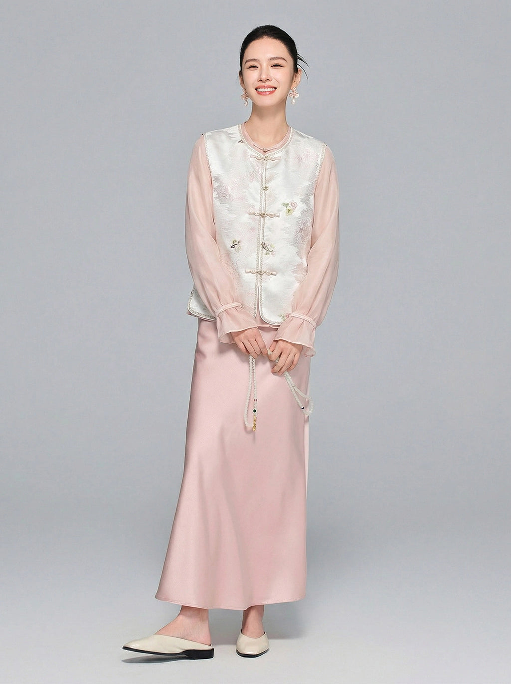 Full body shot of a model wearing a white New Chinese style Tencel vest over a pink sheer blouse and satin midi skirt, showcasing the elegant and graceful aesthetic.