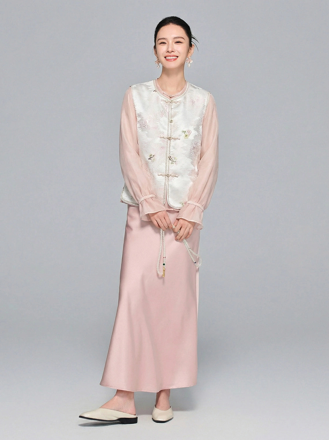 Full body shot of a model wearing a white New Chinese style Tencel vest over a pink sheer blouse and satin midi skirt, showcasing the elegant and graceful aesthetic.
