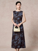 Model wearing a dark brown 100% mulberry silk sleeveless dress with a round neckline and blue floral jacquard pattern, holding a cream handbag.