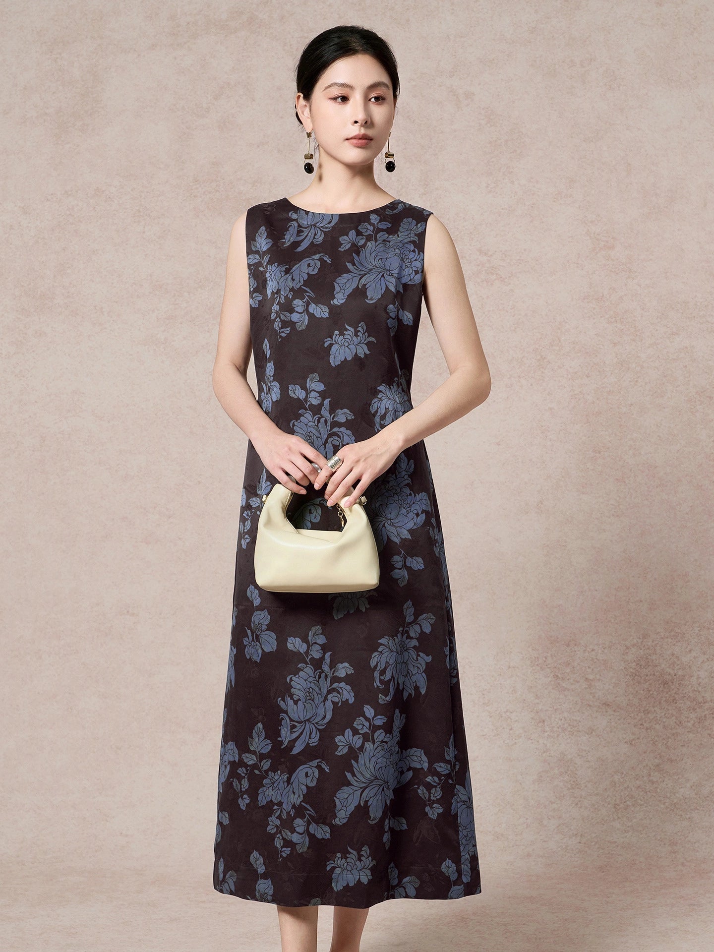 Model wearing a dark brown 100% mulberry silk sleeveless dress with a round neckline and blue floral jacquard pattern, holding a cream handbag.