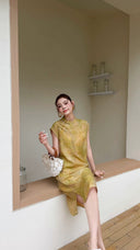 Sitting woman wearing a yellow sleeveless Cheongsam dress with a relaxed fit, paired with a white shell bag and white sandals.