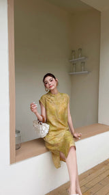 Sitting woman wearing a yellow sleeveless Cheongsam dress with a relaxed fit, paired with a white shell bag and white sandals.