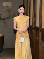 Model wearing a yellow sleeveless Cheongsam dress with beading and a flowing pleated skirt, holding a white handbag, showcasing the elegant drape.