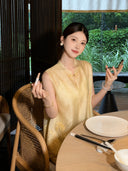 Model sitting at a table wearing a yellow sleeveless Cheongsam vest with gold clasp detail, holding a phone, showcasing the intricate embroidery and relaxed elegance.