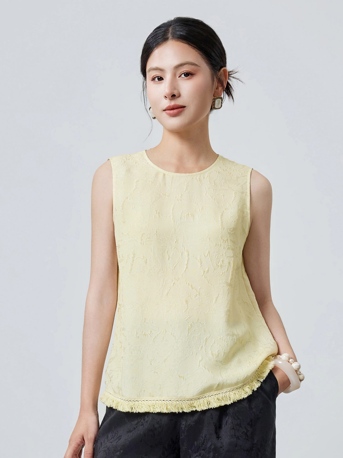 Front view of a cream yellow silk top with a round neckline, textured jacquard pattern, and fringe hem, holding a beige handbag.